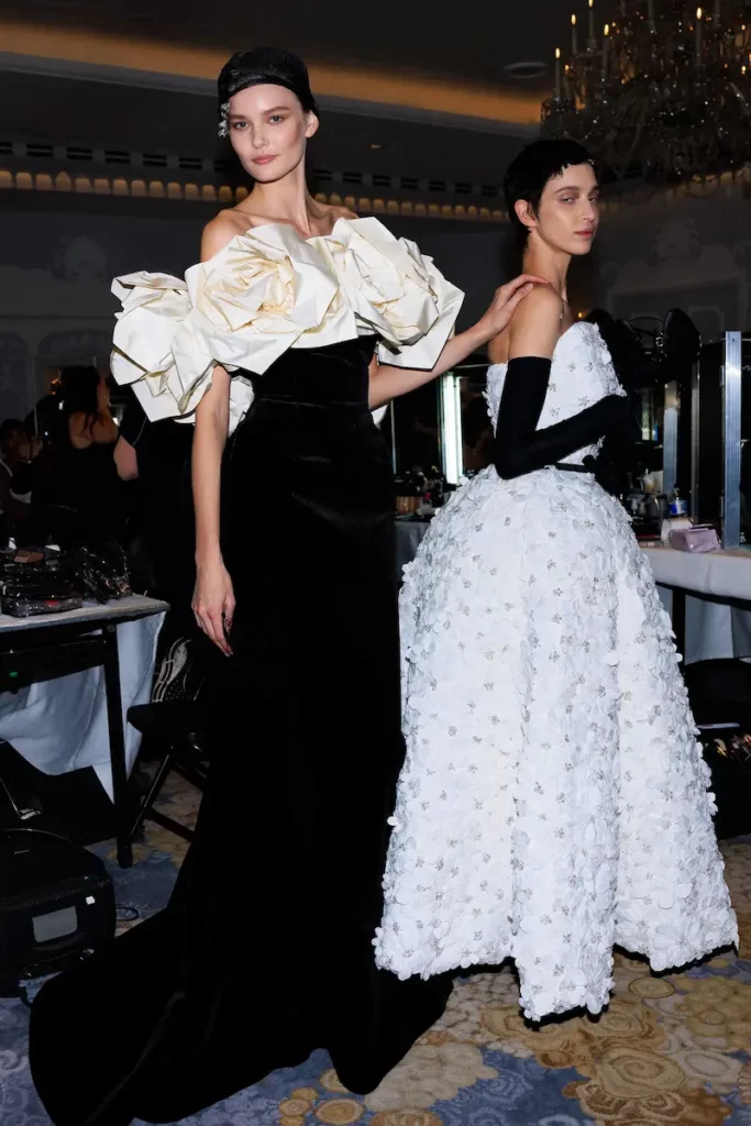 Two women in elgent black-tie gowns, one in black with ruffles, the other in a strapless white dress with gloves.